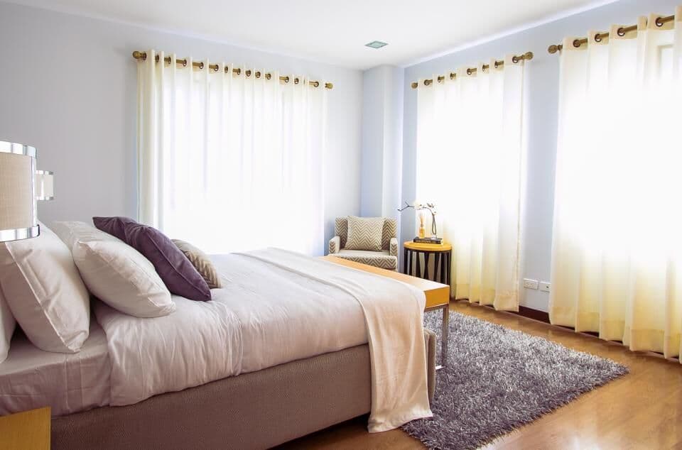 A furnished room with a made bed, curtains and an armchair by the window
